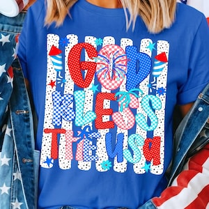 May include: A royal blue t-shirt featuring a graphic print with the text GOD BLESS THE USA. The design includes vertical white stripes with black dots, red and blue patterned letters, firework illustrations, and small star and flower icons.
