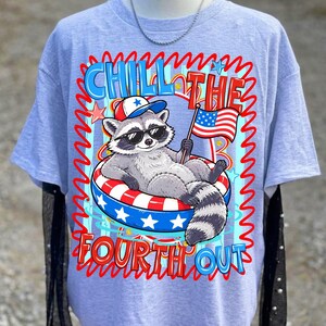 Op de afbeelding: Lichtblauw T-shirt met een cartoon wasbeer met een zonnebril en een patriottische hoed, ontspannen in een rood, wit en blauw zwemband. Het shirt heeft de tekst "Chill the Fourth Out" in een golvend lettertype, met sterren en een Amerikaanse vlag.