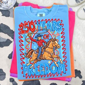 Puede incluir: Una pila de camisetas dobladas en tonos de azul, rosa y naranja. La camiseta superior es azul claro con un gr&aacute;fico de un vaquero montando a caballo, con el texto "250 YEARS OF FREEDOM". Las camisetas est&aacute;n sobre una superficie con estampado de vaca.
