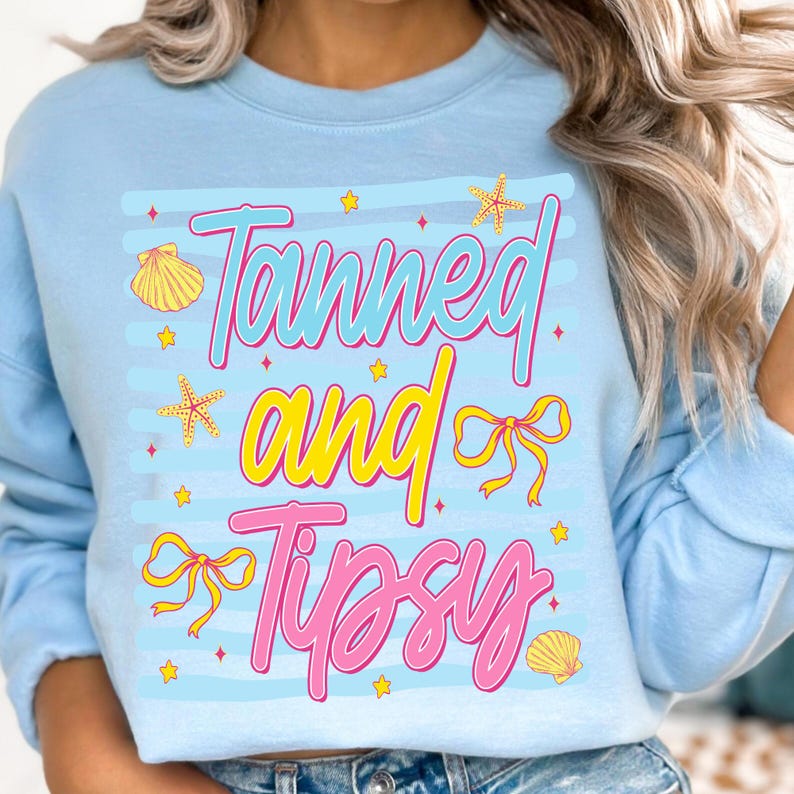 Peut inclure: Sweat-shirt bleu clair avec l'inscription "Tanned and Tipsy" dans une police de caract&egrave;res color&eacute;e et ludique. Le motif comprend des coquillages, des &eacute;toiles de mer, des n&oelig;uds et des &eacute;toiles. Le sweat-shirt a un col rond et des manches longues.