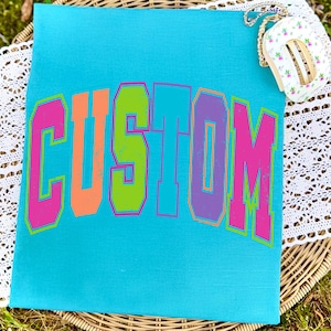 Pu&ograve; includere: Una borsa tote in tela turchese con la scritta "CUSTOM" a grandi lettere multicolore. Le lettere sono delineate in rosa, con arancione, verde e viola che riempiono le lettere. Una piccola borsa floreale con una catena dorata poggia in alto a destra.