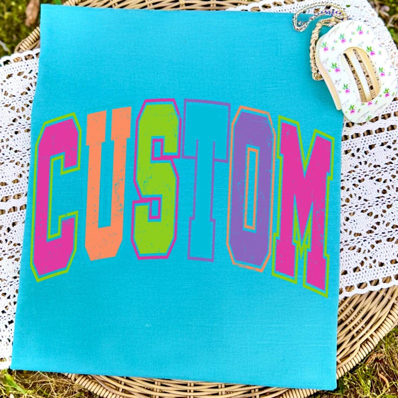 Pu&ograve; includere: Una borsa tote in tela turchese con la scritta "CUSTOM" a grandi lettere multicolore. Le lettere sono delineate in rosa, con arancione, verde e viola che riempiono le lettere. Una piccola borsa floreale con una catena dorata poggia in alto a destra.