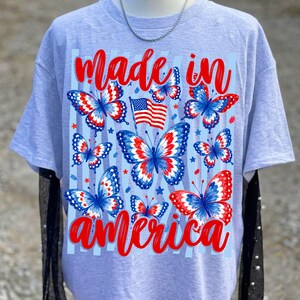 May include: A light grey short-sleeve t-shirt featuring a graphic print of red, white, and blue butterflies, a small American flag, and the red cursive text "made in america" arranged above and below the butterfly and flag design elements.