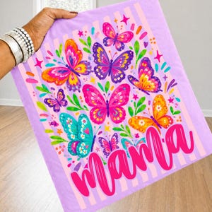 May include: A light purple fabric item featuring a vertical striped pattern and a colorful graphic print of multiple butterflies in shades of pink, orange, yellow, and teal, with the word Mama written in a large, bold, bright pink cursive script.