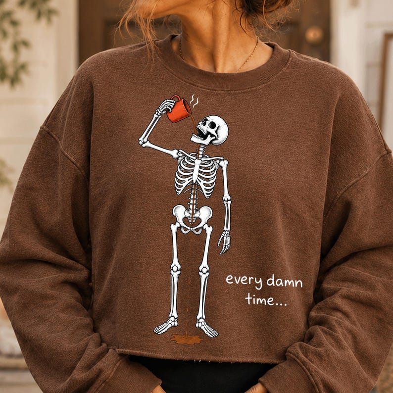 May include: A brown crewneck sweatshirt featuring a graphic of a white skeleton drinking from an orange mug. The coffee spills through the skeleton's ribcage onto the floor. The text "every damn time..." is printed in white next to the skeleton.