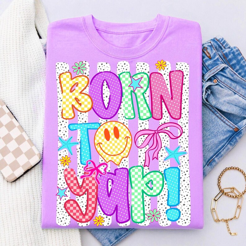 May include: A lilac t-shirt featuring the phrase "BORN TO YAP!" in a vibrant, multi-coloured design. The graphic includes a smiley face, bows, stars, and flowers. The shirt is displayed with blue jeans and a gold bracelet.