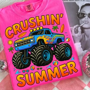 May include: Bright pink t-shirt with the words "CRUSHIN' SUMMER" in orange and yellow. A blue monster truck with flames, palm trees, and beach balls is featured in the design. The shirt is a Comfort Colors brand.