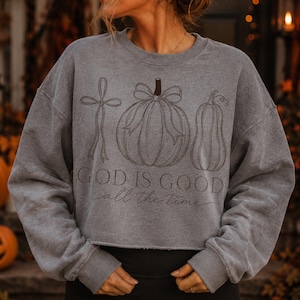 K&ouml;nnte beinhalten: Ein graues Sweatshirt mit Rundhalsausschnitt und einer dunkelbraunen Strichzeichnung, die eine Schleife, einen K&uuml;rbis mit Schleife und einen Flaschenk&uuml;rbis zeigt. Der Text "GOD IS GOOD" ist in fetter Serifenschrift gedruckt, darunter "all the time".