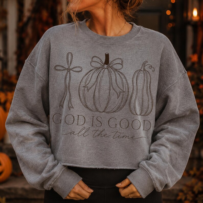 K&ouml;nnte beinhalten: Ein graues Sweatshirt mit Rundhalsausschnitt und einer dunkelbraunen Strichzeichnung, die eine Schleife, einen K&uuml;rbis mit Schleife und einen Flaschenk&uuml;rbis zeigt. Der Text "GOD IS GOOD" ist in fetter Serifenschrift gedruckt, darunter "all the time".
