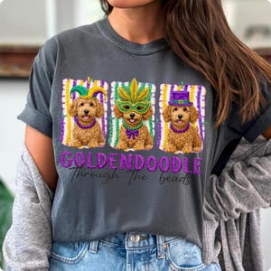 May include: Dark gray t-shirt featuring three Golden Doodle dogs dressed in Mardi Gras attire. Each dog wears a different hat and mask, with the text "GOLDENDOODLE Through The beads" in purple glitter.