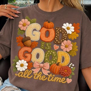 Pu&ograve; includere: Una t-shirt marrone scuro con un design grafico che riporta il testo "GOD IS GOOD all the time". Il design include illustrazioni in stile ricamo di zucche, foglie autunnali, margherite, una ghianda, un fiocco rosa e piccoli cuori.