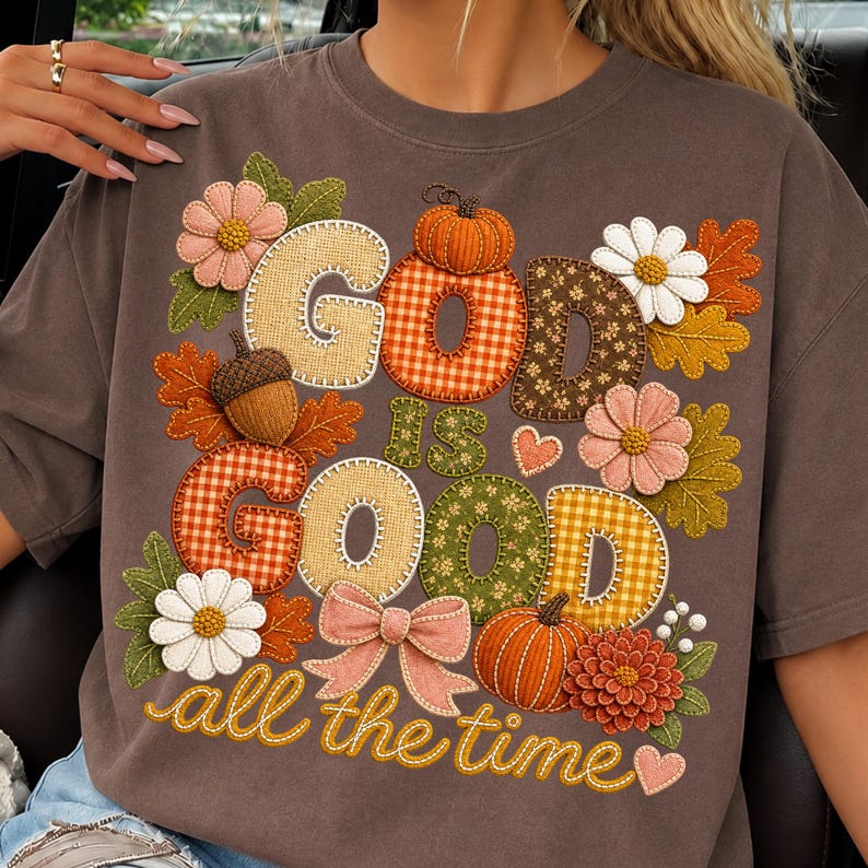 Pu&ograve; includere: Una t-shirt marrone scuro con un design grafico che riporta il testo "GOD IS GOOD all the time". Il design include illustrazioni in stile ricamo di zucche, foglie autunnali, margherite, una ghianda, un fiocco rosa e piccoli cuori.