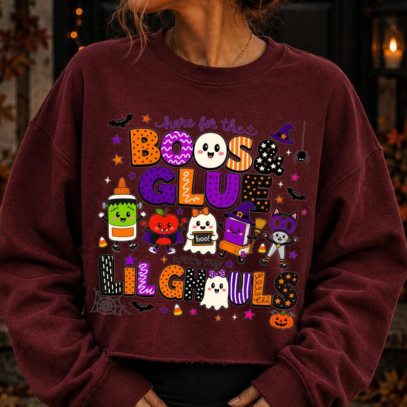 K&ouml;nnte beinhalten: Ein kastanienbraunes Langarm-Sweatshirt mit einer bunten Halloween-Grafik und dem Text "here for the BOOS & GLUE with my LIL GHOULS". Das Design zeigt Zeichentrickfiguren wie eine Klebeflasche, einen Apfel, ein Gespenst, ein Buch und eine Katze.