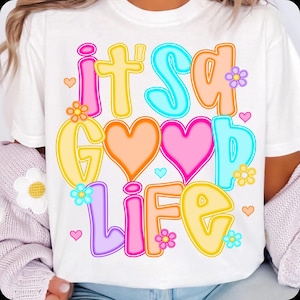 Pode incluir: Camiseta branca com a frase "it's a good life" em letras coloridas. O design inclui cora&ccedil;&otilde;es e flores em rosa, laranja, amarelo, azul e roxo. Uma camiseta gr&aacute;fica casual e alegre.
