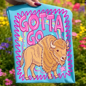 May include: A light blue t-shirt with the words "GOTTA GO" in pink bubble letters, framed by a pink wavy border. A cartoon bison in shades of tan and brown is below the text, with stars and stripes accents.