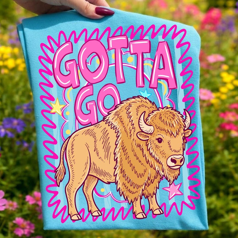 May include: A light blue t-shirt with the words "GOTTA GO" in pink bubble letters, framed by a pink wavy border. A cartoon bison in shades of tan and brown is below the text, with stars and stripes accents.