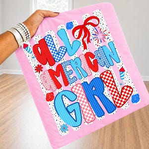 Pode incluir: Uma manta rosa com as palavras "ALL AMERICAN GIRL" em letras vermelhas, brancas e azuis. As letras t&ecirc;m padr&otilde;es xadrez e de bolinhas. A manta tem um la&ccedil;o vermelho, fogos de artif&iacute;cio e detalhes florais.