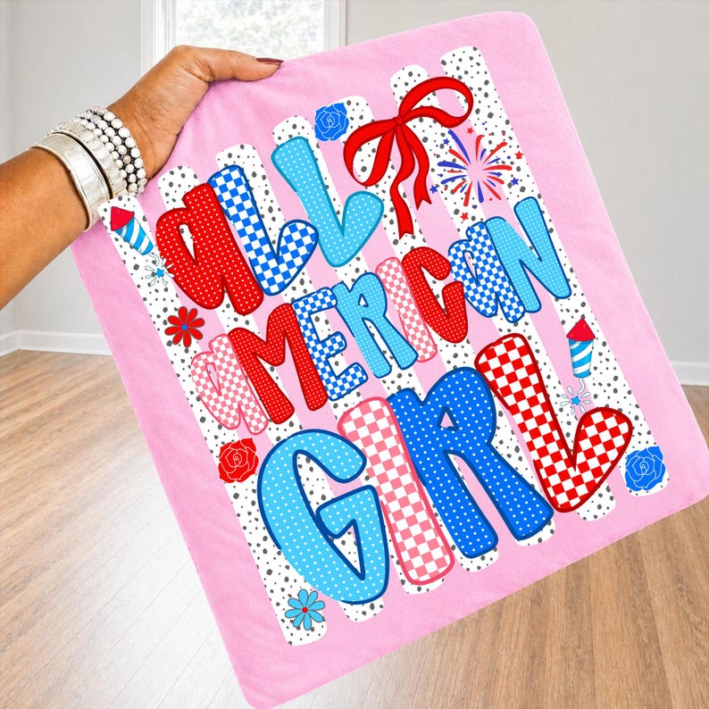 Pode incluir: Uma manta rosa com as palavras "ALL AMERICAN GIRL" em letras vermelhas, brancas e azuis. As letras t&ecirc;m padr&otilde;es xadrez e de bolinhas. A manta tem um la&ccedil;o vermelho, fogos de artif&iacute;cio e detalhes florais.