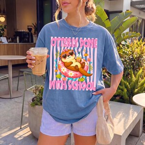 K&ouml;nnte beinhalten: Blaues T-Shirt mit dem Text "Otterly Done With School" und einer Grafik einer Otter mit Sonnenbrille, die auf einem Schwimmring treibt. Die Otter h&auml;lt ein Getr&auml;nk. Das Shirt wird mit gestreiften Shorts getragen.