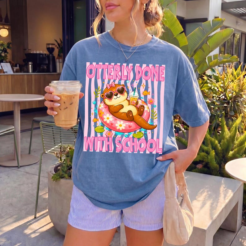 K&ouml;nnte beinhalten: Blaues T-Shirt mit dem Text "Otterly Done With School" und einer Grafik einer Otter mit Sonnenbrille, die auf einem Schwimmring treibt. Die Otter h&auml;lt ein Getr&auml;nk. Das Shirt wird mit gestreiften Shorts getragen.