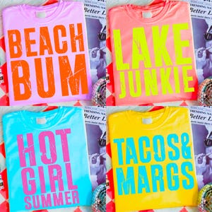 May include: Four colorful t-shirts with bold text: pink with "BEACH BUM", coral with "LAKE JUNKIE", turquoise with "HOT GIRL SUMMER", and yellow with "TACOS & MARGS". Each shirt is a different color and has a unique phrase.