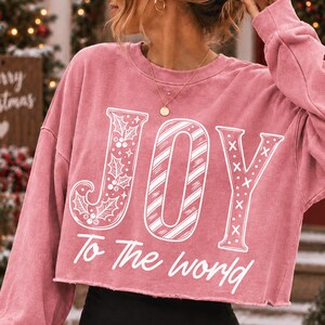 May include: A dusty rose long-sleeve cropped sweatshirt featuring the white graphic text JOY with holly leaves, candy cane stripes, and X patterns, followed by the text To The world in a cursive font across the front of the garment.