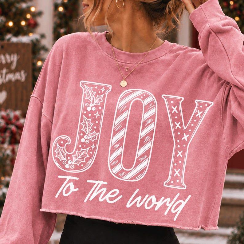 May include: A dusty rose long-sleeve cropped sweatshirt featuring the white graphic text JOY with holly leaves, candy cane stripes, and X patterns, followed by the text To The world in a cursive font across the front of the garment.