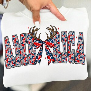 Pu&ograve; includere: Felpa bianca con la scritta "AMERICA" in un motivo mimetico rosso, bianco e blu. Il design include corna marroni e un fiocco. Accenti di stelle rosse sono posizionati intorno alle lettere.