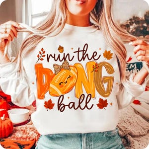 Puede incluir: Una sudadera blanca de manga larga con un estampado gr&aacute;fico que incluye el texto "run the ball". El dise&ntilde;o presenta un bal&oacute;n de f&uacute;tbol americano naranja con un lazo de estampado de leopardo, hojas de oto&ntilde;o, bayas y una bellota.
