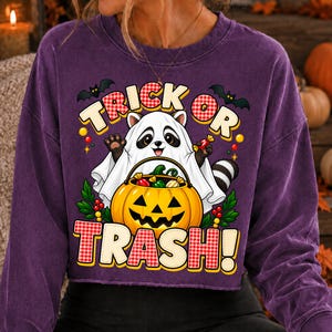 May include: A purple long-sleeve sweatshirt featuring a graphic of a raccoon wearing a white ghost sheet and holding a jack-o'-lantern bucket filled with sweets. The text reads TRICK OR TRASH! with bats and red berries surrounding the design.