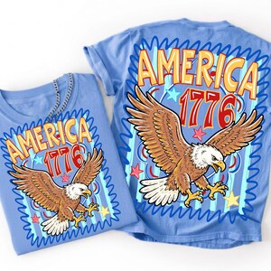 May include: Two light blue t-shirts with a patriotic design. Each shirt features the word "AMERICA" in orange and yellow, the number "1776" in red, and a brown eagle with outstretched wings. The design is framed by blue wavy lines and stars.