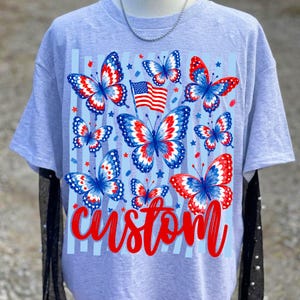 Peut inclure: Un t-shirt gris clair &agrave; manches courtes avec une impression graphique de plusieurs papillons aux motifs rouges, blancs et bleus. Un petit drapeau am&eacute;ricain est plac&eacute; parmi les papillons et le mot custom est &eacute;crit en &eacute;criture rouge en bas.