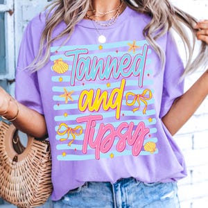 Peut inclure: Un t-shirt lilas avec l'inscription "Tanned and Tipsy" dans un design color&eacute;. Le texte est plac&eacute; sur un fond ray&eacute; bleu clair, orn&eacute; de coquillages et d'&eacute;toiles de mer. Le t-shirt a une coupe d&eacute;contract&eacute;e.