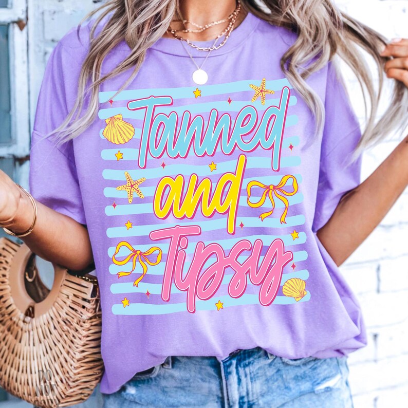 Peut inclure: Un t-shirt lilas avec l'inscription "Tanned and Tipsy" dans un design color&eacute;. Le texte est plac&eacute; sur un fond ray&eacute; bleu clair, orn&eacute; de coquillages et d'&eacute;toiles de mer. Le t-shirt a une coupe d&eacute;contract&eacute;e.