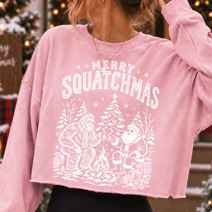 K&ouml;nnte beinhalten: Ein hellrosa Langarm-Sweatshirt mit offenem Saum und einem wei&szlig;en Grafikdruck, der Bigfoot und den Weihnachtsmann an einem Lagerfeuer zeigt. &Uuml;ber der Illustration mit Tannenb&auml;umen und Geschenken steht der Text MERRY SQUATCHMAS.