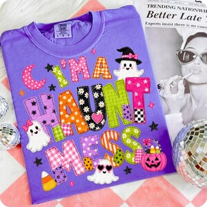 May include: A purple t-shirt featuring the text "I'M A HAUNT MESS" in colorful, patterned block letters. The design includes three cartoon ghosts, a pink jack-o'-lantern bucket filled with candy corn, and small black stars on a purple background.