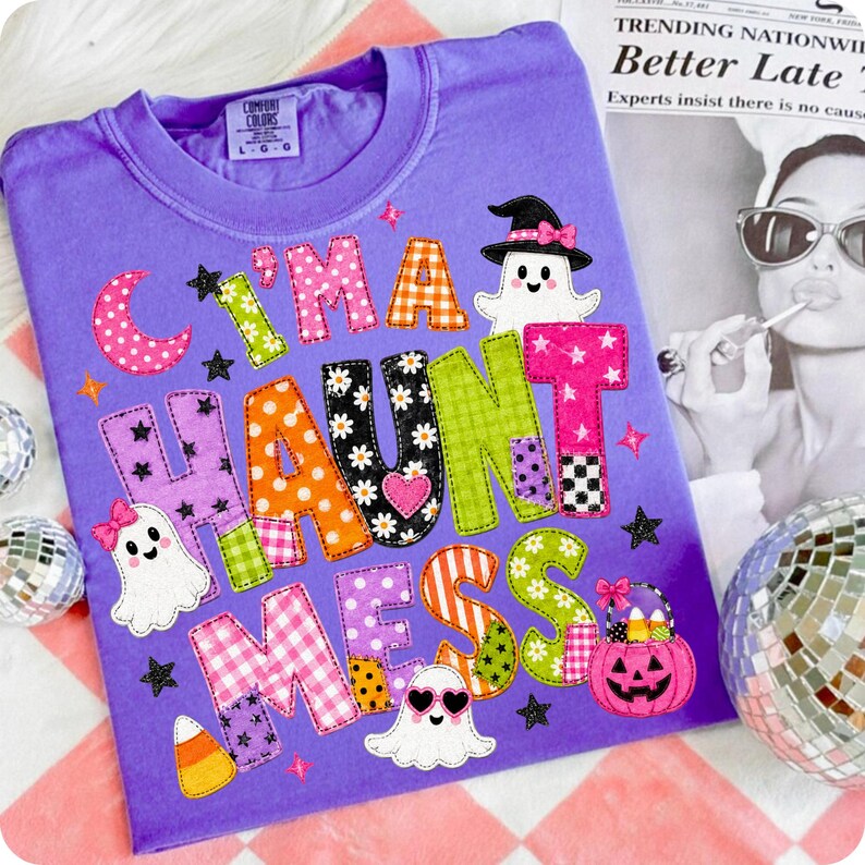 May include: A purple t-shirt featuring the text "I'M A HAUNT MESS" in colorful, patterned block letters. The design includes three cartoon ghosts, a pink jack-o'-lantern bucket filled with candy corn, and small black stars on a purple background.