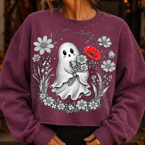 K&ouml;nnte beinhalten: Ein dunkelweinrotes Langarm-Sweatshirt mit einer zentralen Grafik eines wei&szlig;en Geistes, der einen Blumenstrau&szlig; h&auml;lt. Das Design umfasst schwarz-wei&szlig;e florale Linienzeichnungen, die den Geist umgeben, sowie eine einzelne leuchtend rote Mohnblume.