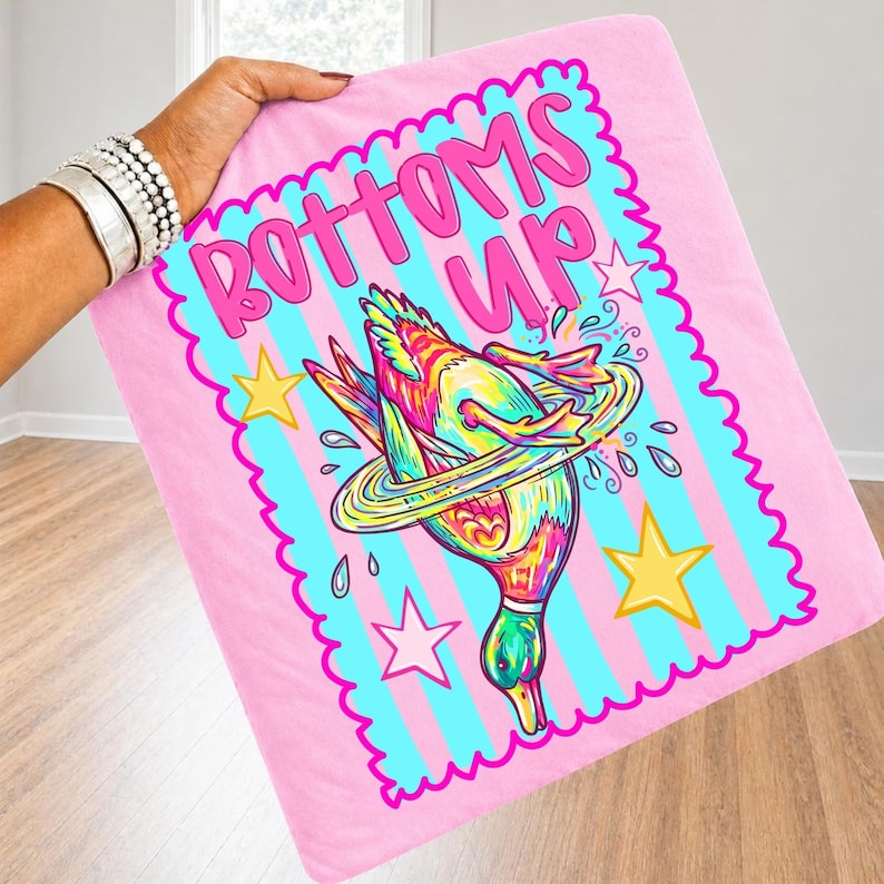 May include: Pink blanket featuring a cartoon duck illustration and the words "BOTTOMS UP" in pink. The duck is colourful with a ring around it, and there are stars and water droplets. The blanket is held up by a person.