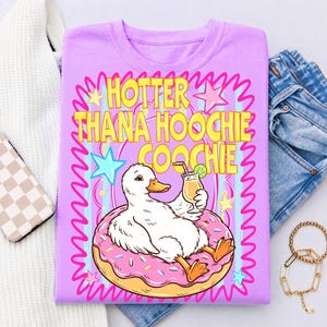 Może przedstawiać: Liliowa koszulka z kaczorem z kresk&oacute;wki na r&oacute;żowym pączku, trzymającym koktajl. Tekst "HOTTER THANA HOOCHIE COOCHIE" jest w kolorze ż&oacute;łtym i r&oacute;żowym. Projekt zawiera gwiazdy i faliste linie.