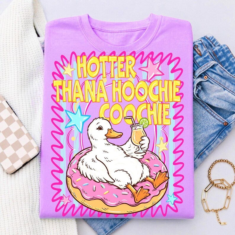 Może przedstawiać: Liliowa koszulka z kaczorem z kresk&oacute;wki na r&oacute;żowym pączku, trzymającym koktajl. Tekst "HOTTER THANA HOOCHIE COOCHIE" jest w kolorze ż&oacute;łtym i r&oacute;żowym. Projekt zawiera gwiazdy i faliste linie.