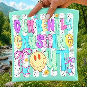 Può includere: Una borsa tote verde menta con la scritta "CURRENTLY CRASHING OUT" in lettere a bolle colorate. Il design include una faccina sorridente, fiocchi, stelle e fiori. Lo sfondo presenta strisce bianche e pois neri.