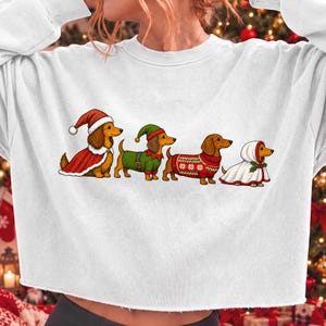 May include: A white long-sleeve cropped sweatshirt featuring a graphic print of four brown dachshunds. The dogs are dressed in holiday attire, including a Santa suit, an elf costume, a red patterned jumper, and a white hooded winter cloak.