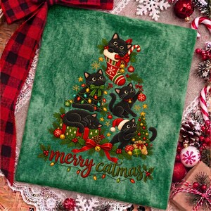 K&ouml;nnte beinhalten: Ein gr&uuml;ner Stoffaufn&auml;her mit einer Grafik aus f&uuml;nf schwarzen Katzen in Tannenbaumform, verziert mit Weihnachtskugeln, Lichterketten und einem roten Weihnachtsstrumpf. Der Text "merry catmas" steht in roter Schreibschrift unten.