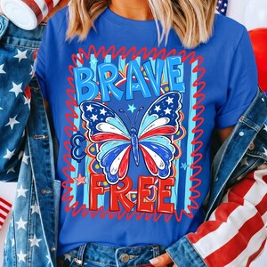 May include: A blue t-shirt featuring the words "BRAVE FREE" in a red and blue design. A butterfly with red, white, and blue wings is in the centre. The shirt has a patriotic theme.