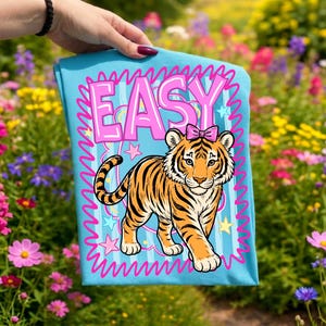 May include: A light blue t-shirt with a cartoon tiger wearing a pink bow, set within a pink wavy border and the word "EASY". The tiger has orange and black stripes. The background is a vibrant garden filled with flowers.