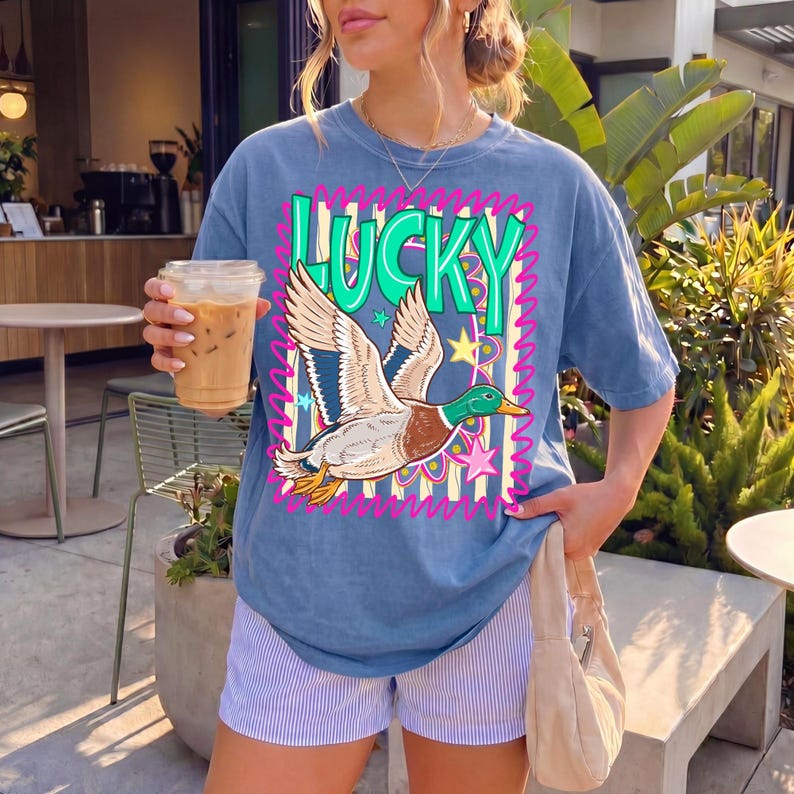 May include: Blue t-shirt with the word "LUCKY" in green and pink, featuring a duck graphic. The shirt is paired with striped shorts. The person is holding a beverage in a clear cup.
