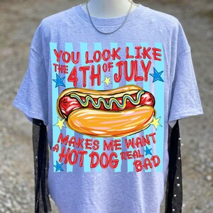 May include: A light gray t-shirt with a graphic of a hot dog and the text "YOU LOOK LIKE THE 4TH OF JULY MAKES ME WANT A HOT DOG REAL BAD". The hot dog is in a bun with mustard.
