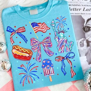 K&ouml;nnte beinhalten: Hellblaues T-Shirt mit patriotischen Illustrationen. Das Design umfasst eine wehende amerikanische Flagge, die Freiheitsstatue, Feuerwerk, Schleifen, einen Hotdog und ein Eis am Stiel, alles in Rot, Wei&szlig; und Blau.