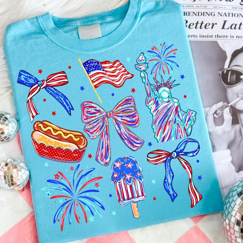 K&ouml;nnte beinhalten: Hellblaues T-Shirt mit patriotischen Illustrationen. Das Design umfasst eine wehende amerikanische Flagge, die Freiheitsstatue, Feuerwerk, Schleifen, einen Hotdog und ein Eis am Stiel, alles in Rot, Wei&szlig; und Blau.